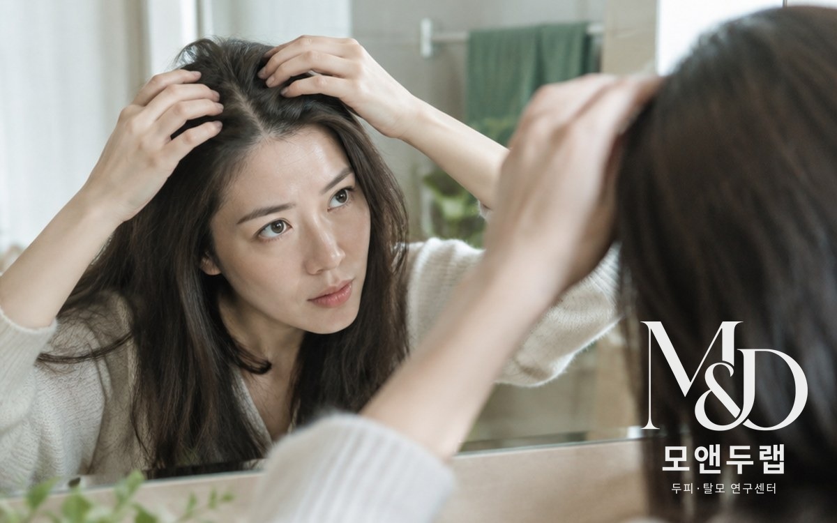 거울 앞에서 양손으로 머리를 들어 올려 두피 상태를 살펴보는 여성의 모습